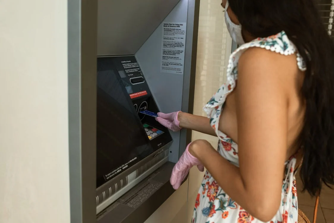 Kobieta w maseczce i rękawiczkach korzysta z bankomatu wewnątrz budynku.